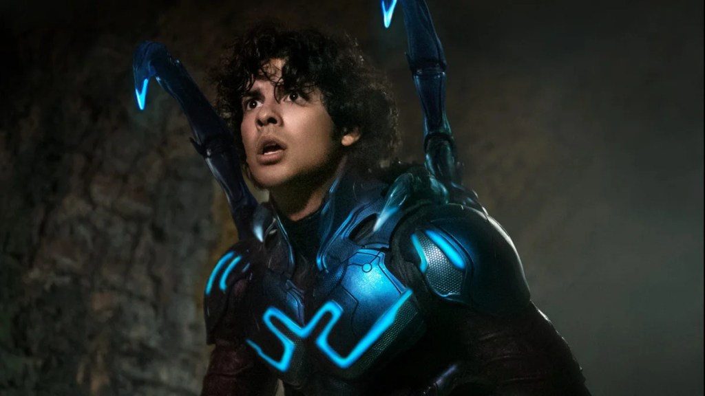 Un jeune homme latino porte un costume de super-héros bleu fluo, le bleu éclairé, avec son casque baissé, montrant son visage. Deux antennes sortent de ses épaules.