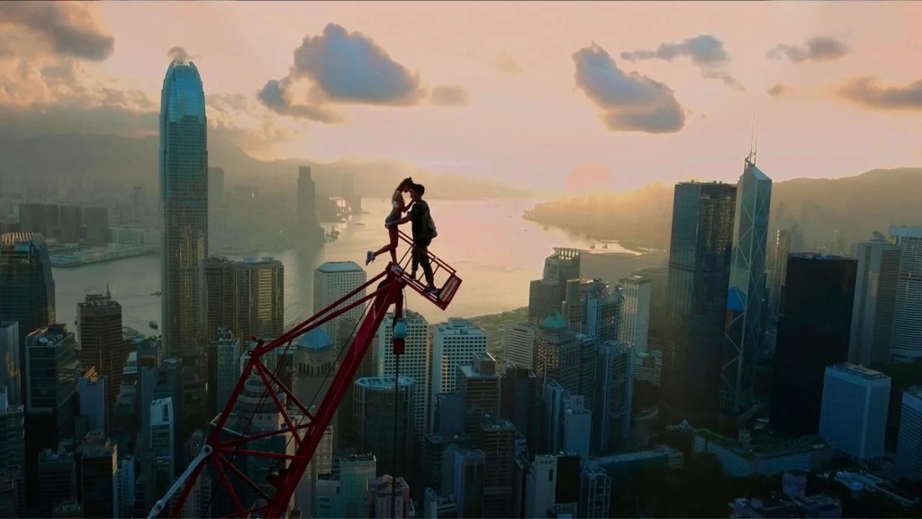 Un homme et une femme partagent un moment d'intimité au sommet d'une grue, au-dessus d'un paysage urbain, le soleil illuminant le ciel en arrière-plan depuis l'horizon.
