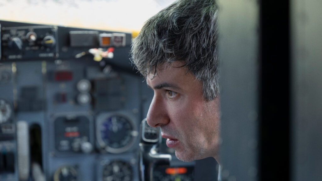 Nathan Fielder est assis dans le cockpit d'un avion, regardant avec une concentration intense. L'arrière-plan montre divers instruments et commandes d'avion, mettant l'accent sur le réglage. Son expression est sérieuse et contemplative, suggérant un moment élevé ou chargé émotionnellement.