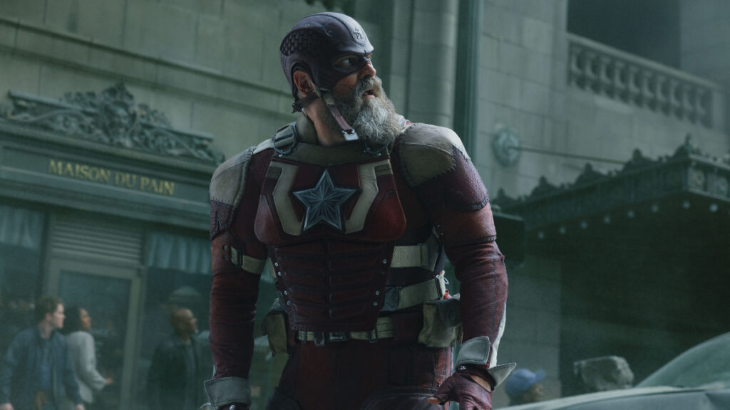 Un homme musclé dans un costume blindé rouge et gris se tient alerte dans un cadre urbain, portant un casque avec un emblème d'étoile et une barbe grise pleine. Son uniforme, marqué par une grande étoile sur la poitrine et les ceintures utilitaires à sa taille, ressemble à un super-soldat sur le thème patriotique.