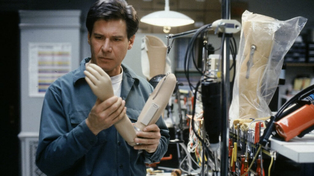Harrison Ford, en tant que Dr Richard Kimble dans le fugitif, examine un bras prothétique dans un laboratoire encombré rempli de fils, d'outils et de membres de mannequin. Il porte une expression sérieuse et une chemise de travail bleue alors qu'il étudie un indice clé dans le thriller plein de suspense.