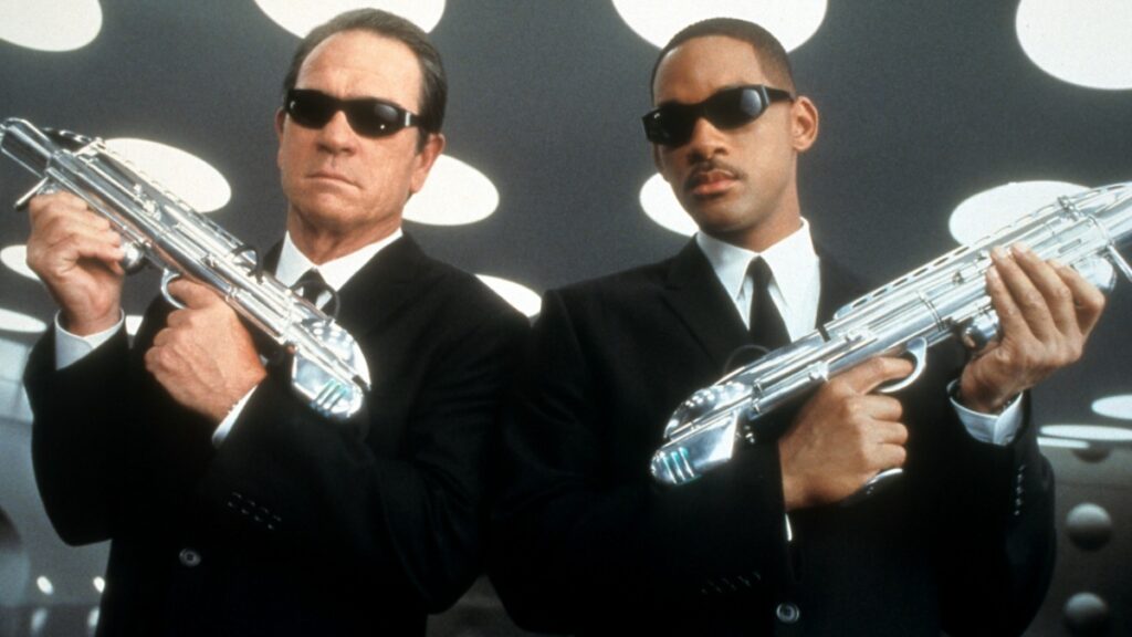 Deux hommes vêtus de costumes noirs élégants et de lunettes de soleil sombres se tiennent en toute confiance avec un paysage urbain futuriste derrière eux. L'un détient un appareil de neuralyzer en argent, tandis que les deux dégagent un air de mystère et d'autorité. Leur look emblématique suggère qu'ils sont des agents de la franchise "Men in Black"