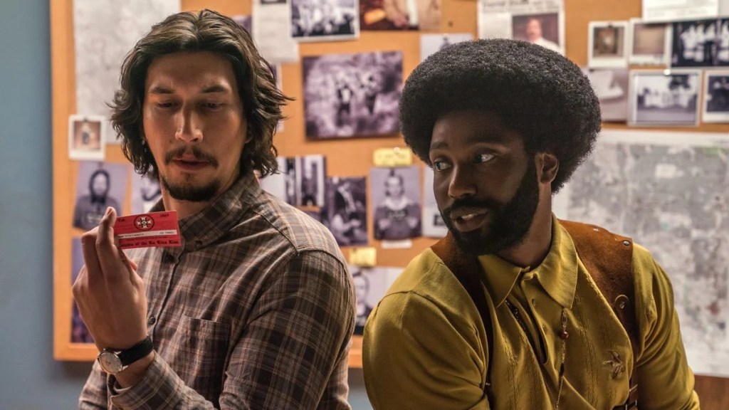 Deux hommes debout devant un babillard recouvert de photos et de notes. L'homme de gauche a de longs cheveux noirs, une barbe, et porte une chemise à carreaux tout en tenant un carton rouge. L'homme à droite a un afro, une barbe, et porte une chemise jaune avec un gilet.