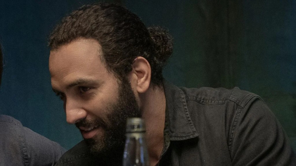 Un homme avec une barbe et des cheveux tirés se penche en avant en milieu de conversation, souriant légèrement