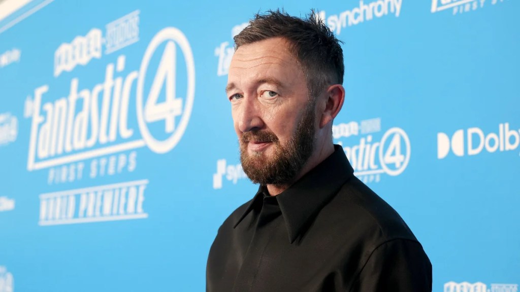 Ralph Ineson sourit à la caméra sur le tapis rouge "Fantastic Four: First Steps" avec une toile de fond de logo bleu derrière lui