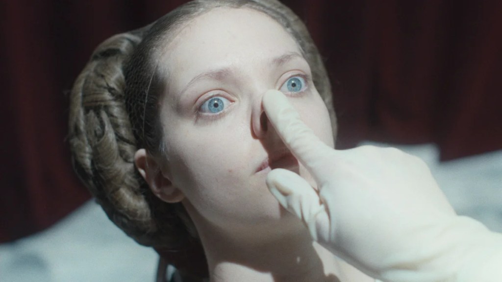 Une femme aux grands yeux bleus perçants est allongée tandis qu’une main gantée appuie doucement sur son nez. Ses cheveux sont coiffés en boucles complexes de chaque côté de sa tête, évoquant un look vintage ou théâtral. L'éclairage est intense, donnant à la scène un ton surréaliste et clinique, tandis que des rideaux rouges profonds sont flous en arrière-plan.